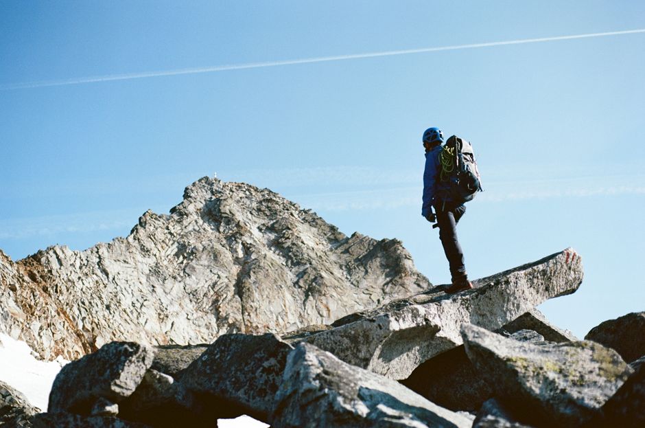 Full body of anonymous person on rocky rough formation among mountains under bright blue sky with airplane cloud